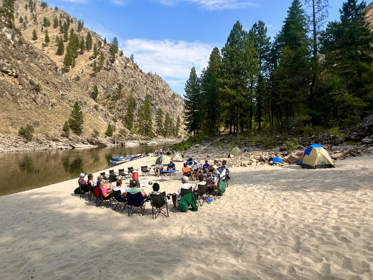 The group seated in a circle on a sandy beach by the river